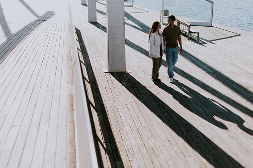 Young couple walks hand in hand, casting long shadows on the wooden boardwalk by the sparkling sea in Barcelona