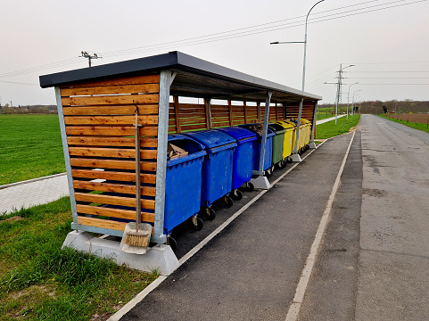 pergola roof with bus stop. combined with bin sorting. plastic, paper, glass separated into colored bins. wooden slats, blue sky, clouds, broom, trowel, sidewalk, street