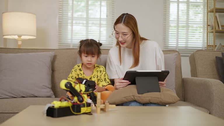 Small young girl using a wired remote control to move her robotic arm toy around while her older sister sitting beside her with a computer tablet guiding and teaching her