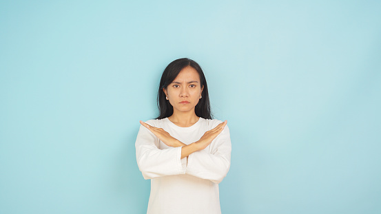 Asian lady in white showing rejection with arms crossed, a sign of 'no', on light blue backdrop