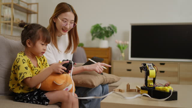 Small young girl using a wired remote control to move her robotic arm toy around while her older sister sitting beside her with a computer tablet guiding and teaching her