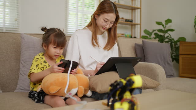 Small young girl using a wired remote control to move her robotic arm toy around while her older sister sitting beside her with a computer tablet working