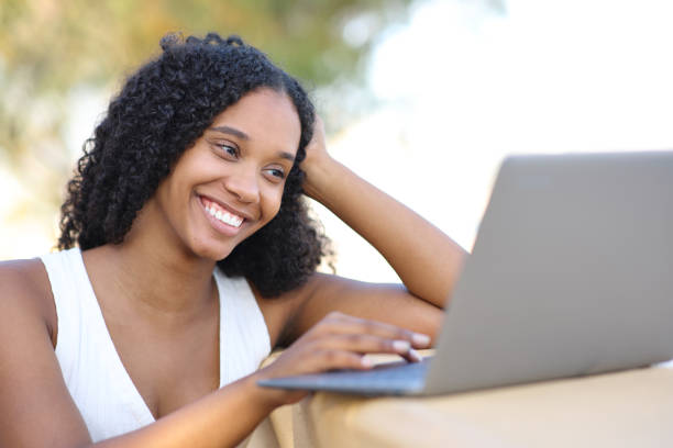 istock Happy black woman using laptop watching online content outdoors 2147517036