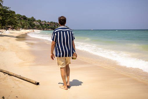 Man enjoys a leisurely walk along a beautiful sandy beach, holding a coconut drink at Ko Phangan Island, Thailand.