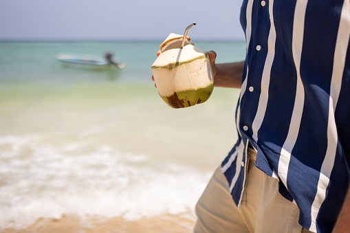 Unrecognizable man enjoys a leisurely walk along a beautiful sandy beach, holding a coconut drink at Ko Phangan Island, Thailand.
