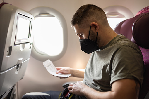 Man wearing a black face mask, is looking at a ticket or boarding pass while seated on an airplane.