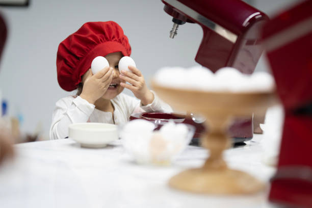 Little girl puts 2 eggs on her eyes and looks at the camera with a smile. There is a red dough making machine in front of them. There's an egg container on the counter. Preparing dough
