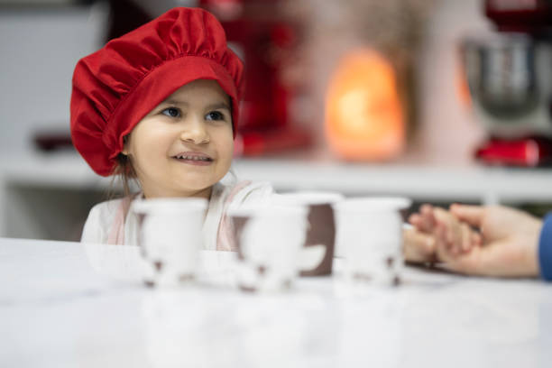 Little girl in pink kitchen apron and red chef hat is sitting at the kitchen counter and holding her mother's hand