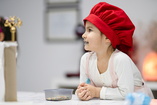 Little girl in pink kitchen apron and red chef hat is smiling