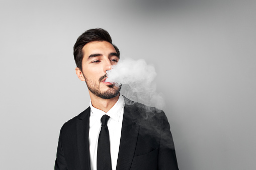 Man businessman in suit smokes e-cigarette and blows steam from his mouth, smoking and health stress in gray background close-up of face at wide camera angle. High quality photo