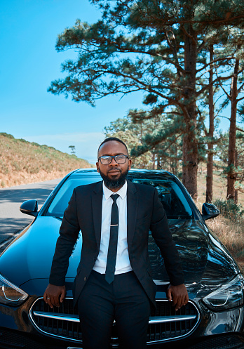 An elegant man in a sharp suit poses confidently in front of his high-end vehicle, surrounded by a serene forest landscape, exuding success and sophistication.