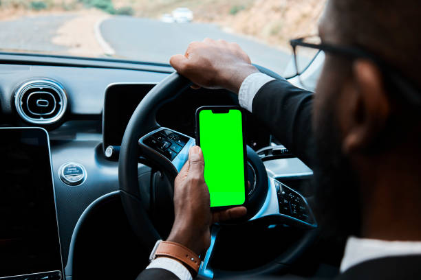 Close-up view of a male professional in formal wear navigating a car while using a smartphone with a replaceable green screen. The scene hints at multitasking, urban commuting, and technology in motion.