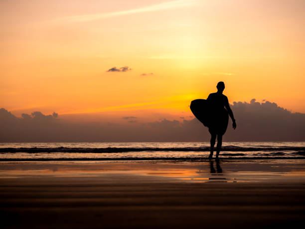 istock Man silhouette holding surfing board, walking on dark seashore with copy space. Surfer standing on sunset sky over the sea water background. Summer sand beach in evening time. dusk seascape scene. 2147522803