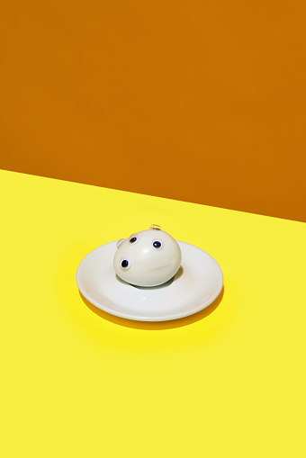 Boiled egg with googly eyes on it in small round plate lying on yellow orange background. Artistic food representation. Concept of food pop art photography, creativity, quirky style