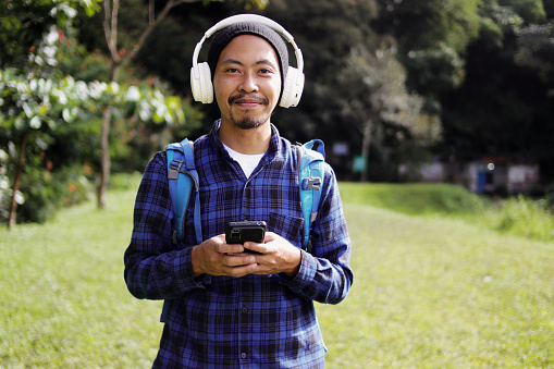Smiling young Asian man in a casual outfit, wearing a beanie, plaid shirt, backpack, and headphones, holds his phone and looks directly at the camera while enjoying a morning walk on a nature trail.