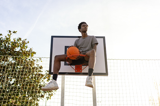 Low angle view portrait of a young cool african american young man sitting on a basketball hoop looking away