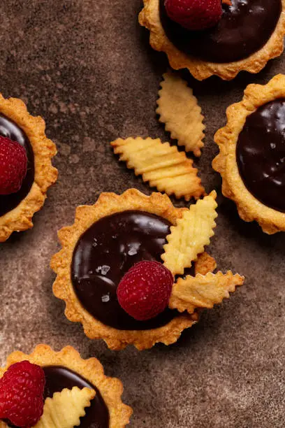 Vertical image of Salted Chocolate Mini Tarts with chocolate ganache cream and raspberry with foodstyling,  on a brown table surface.