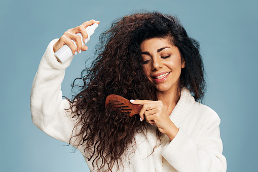 Hairstyling routine. Enjoyed tanned curly Latin lady in bathrobe Spraying On Hair For Repair split ends posing isolated on pastel blue background, using hairbrush, smiling. Hair Care Ad Concept