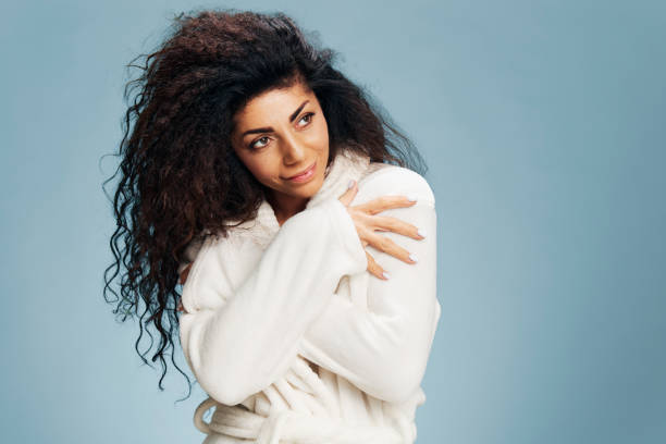 istock Pensive tanned cheerful curly Latin lady touch soft warm bathrobe, hugging herself, smiling, looking aside posing isolated on pastel blue background. Natural cosmetics, self care concept. Copy space 2147532880