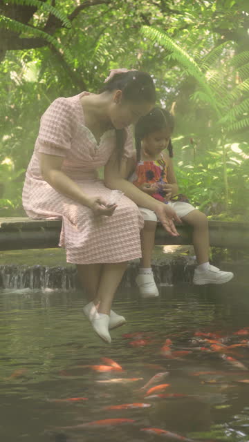 Thai mother and daughter feed koi fish in a shady, misty garden.