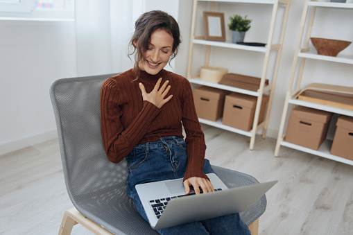 Cheerful smiling happy curly beautiful lady enjoy accepts congratulations from colleagues on video call conference using laptop loves her work sitting in chair at home. Modern Profession Remote Job