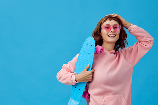 Cute smiling pretty redhead lady in pink hoodie sunglasses with penny board look at camera posing isolated on blue studio background. Copy space Banner Offer. Fashion Cinema. Holiday activity