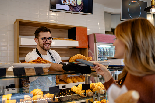 An image showing a woman making a payment with her mobile phone in a bakery, exemplifying the ease of contactless, tech-savvy purchasing methods