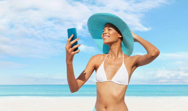 istock Happy young woman at the beach side using mobile phone, wearing a turquoise sun hat and bikini, portrait of African latin American woman in sunny summer day with blue sky, concept of a summer holiday 2147538708