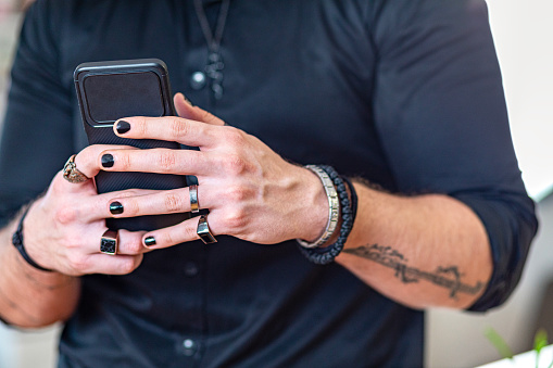 Tattooed arms and painted fingernails of a muscular man. Young adult man using smart phone to pay bill, to make online payment service or online shopping. Cashless payment using mobile application and make money transfer completely via internet.