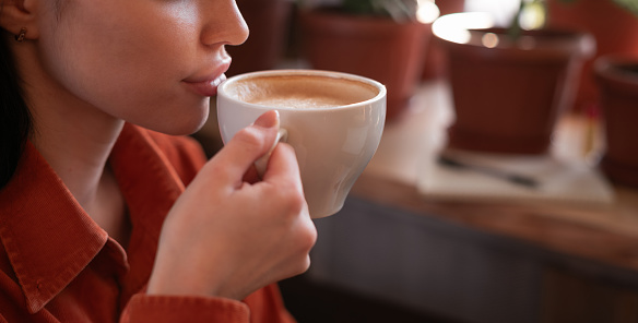 Charming young brunette woman enjoying aroma coffee in cafe holding cup with latte near her lips. Banner. Copy space