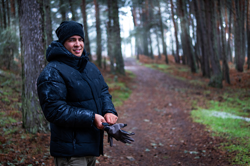 Latino hiker, clad in mountain attire, grinning amidst snow-dusted pines, exploring Madrid's Sierra.