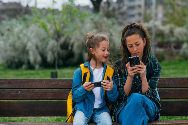 istock Little boy accompanied by his older sister using smart phone sitting on a bench. 2147541671