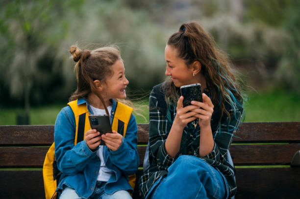 istock Little boy accompanied by his older sister using smart phone sitting on a bench. 2147541695
