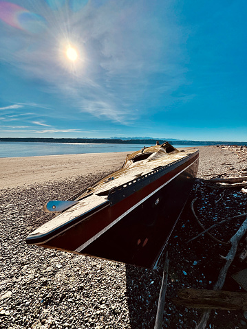 A beached, weathered sailboat lies on its hull atop rocks and seashells on a Hood Canal beach at low tide.  Driftwood lines the high tide level on the beach and the Olympic Mountains can be seen in the distance.  The sun shines brightly overhead on a sunny day in Puget Sound.