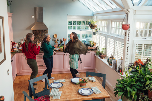 A wide shot of a group of mature female friends gathered together inside a home in Hexham, Northumberland. They are in the kitchen, preparing for a meal together, dancing and holding wineglasses and enjoying time together.