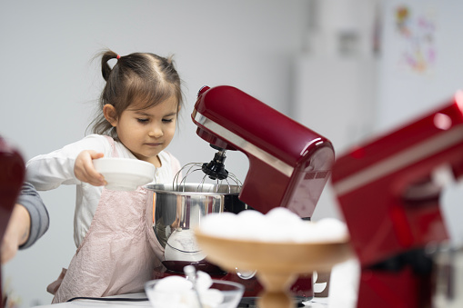 Little cute girl is pouring ingredients into the mixing device. There is a red dough making machine in front of them. There's an egg container on the counter. Preparing dough