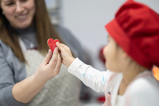 Little girl in pink kitchen apron and red chef hat receives a red heart-shaped cookie from her teacher. They are smiling