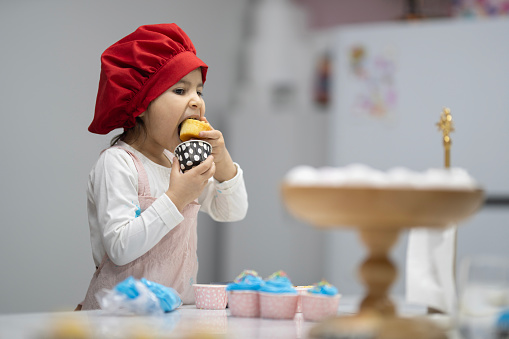 While the little girl was preparing the cupcakes, she couldn't resist taking one of them to her mouth and biting it big. Little girl in pink kitchen apron and red chef hat is happy