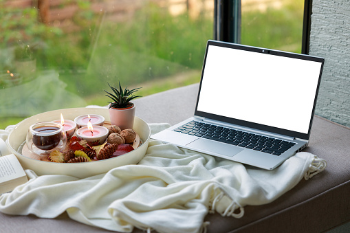 A comfortable working environment at home. Laptop, coffee and burning candles on the window sill. Blank screen with convenient copy space for adding your own design elements