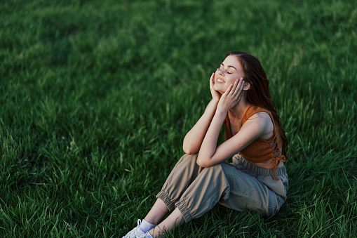 A beautiful girl relaxing in nature sitting in pants and top on the green grass in the rays of the setting summer sun. The concept of health and care for the body and the environment . High quality photo