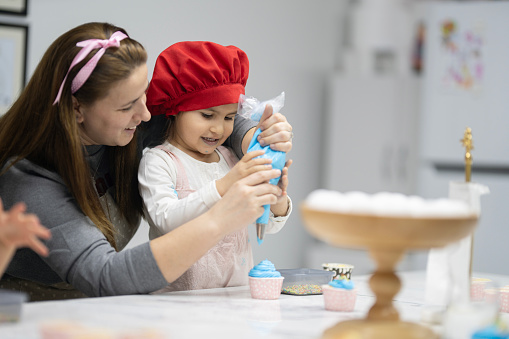Teacher helps little girl pour whipped cream on cupcake. Little girl in pink kitchen apron and red chef hat is smiling