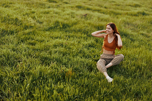 The sunset light of a summer evening illuminates a beautiful young redheaded woman sitting on the grass in green pants and an orange top and smiling at the sun. High quality photo