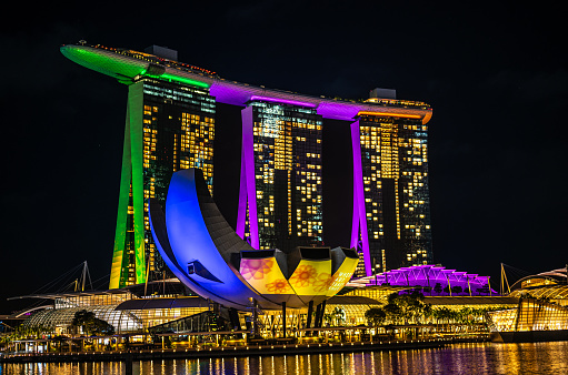 Marina Bay, Singapore - February, 10 2023: Stock photo showing tourists and locals walking on Marina Bay promenade with a view across Singapore waterfront of the Marina Bay Sands hotel and SkyPark illuminated at night.