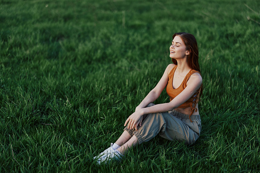 A happy woman looks out at the setting summer sun sitting on the fresh green grass in the park and smiling, view from above. The concept of self-care. High quality photo