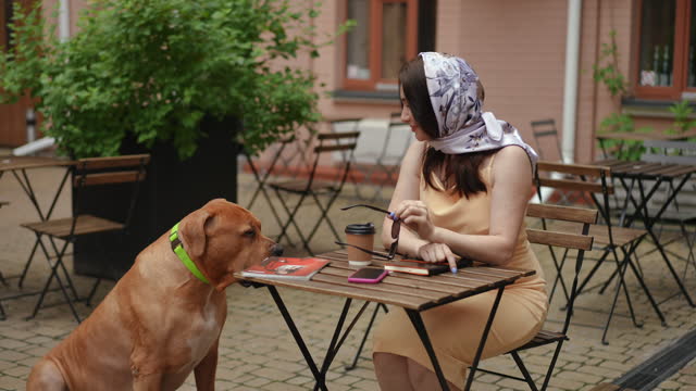 Slow motion. Stylish woman takes off her sunglasses and looks at her dog with a smile while sitting at a table outdoors with a plastic cup of coffee.