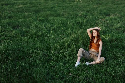A girl in comfortable clothes sat on the grass after a walk in the park to meditate and rest in the sunlight. High quality photo