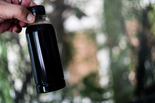 A hand holding freshly black coffee in bottle on blurry nature background.