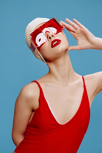 Summer fashion shooting. A sophisticated red-lipped lady in a swimming cap and a red swimsuit touches her glasses and looks up posing on a blue background. High quality photo