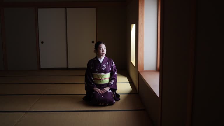 Female tourist in purple kimono sitting on heels in dark Japanese tatami room and bowing