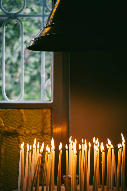 Lit prayer candles on a candlestand at a serene chapel during evening hours.In the background, a metal grid window and a church bell on the top right corner